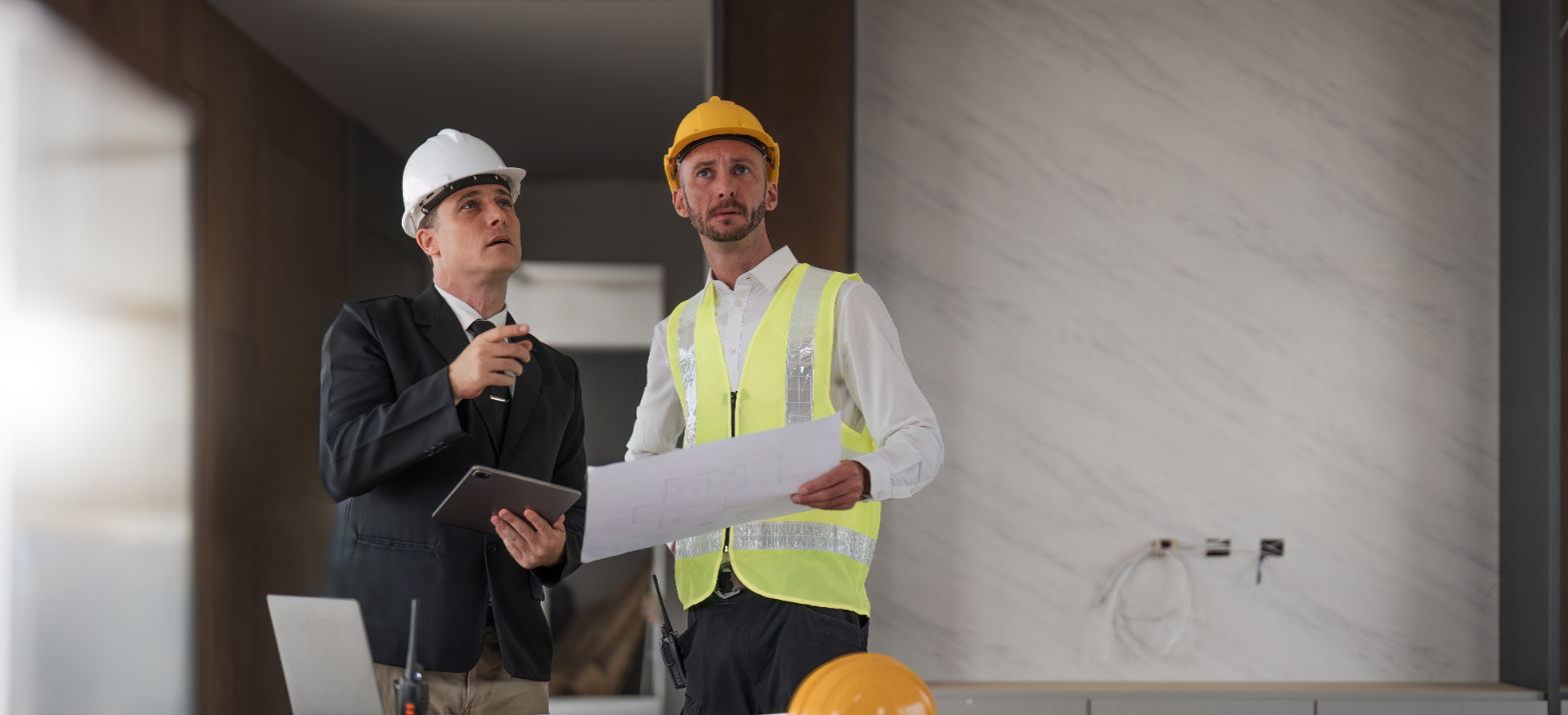 Two men wearing hard hats compare documents in a commercial building during construction